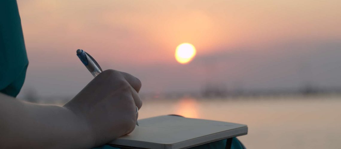 Close up view of the hand of a woman writing in her diary at sunset with the glowing orb of the sun reflected over a still ocean